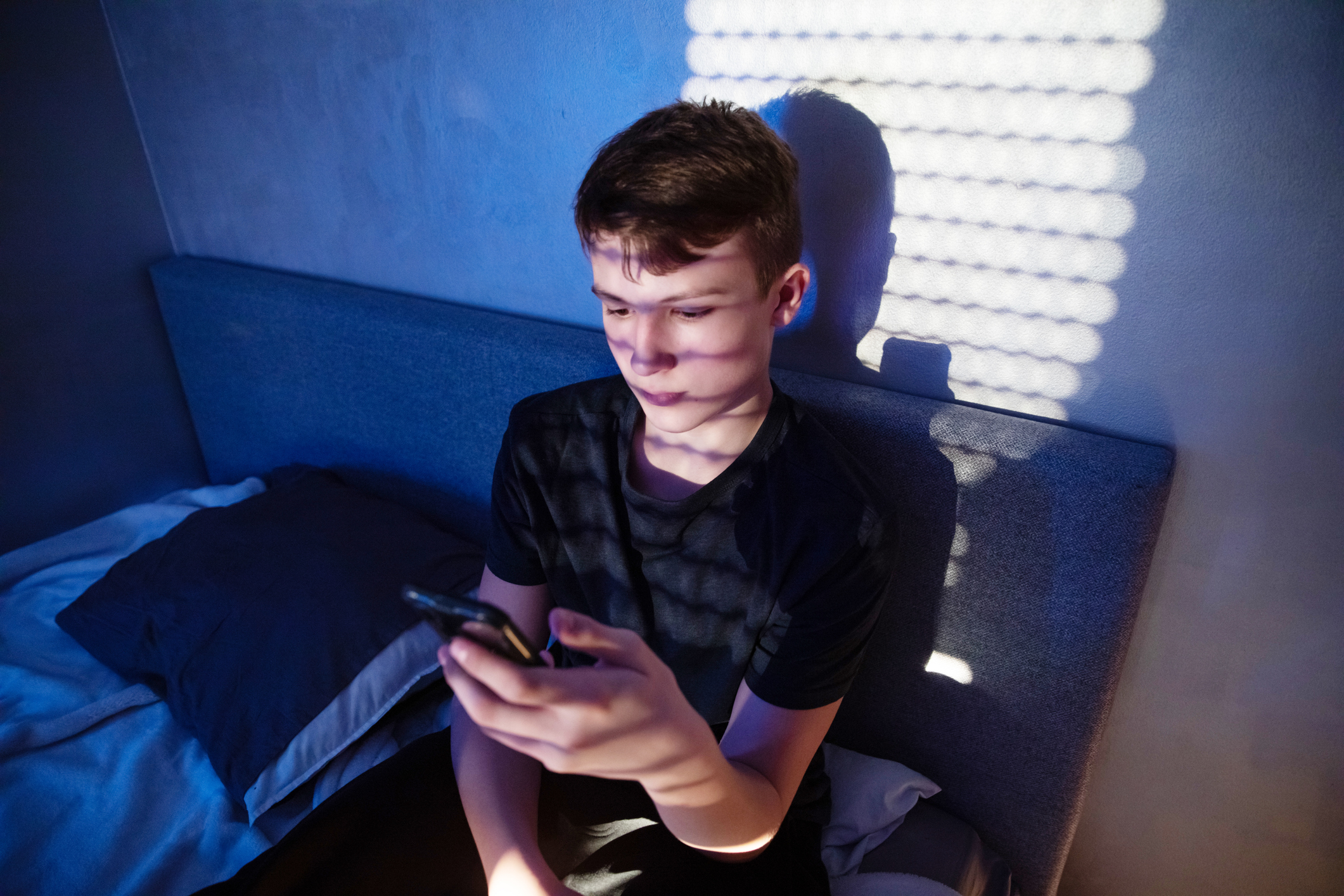 A young boy sits on his bed in a dimly lit room. He is looking at his phone.