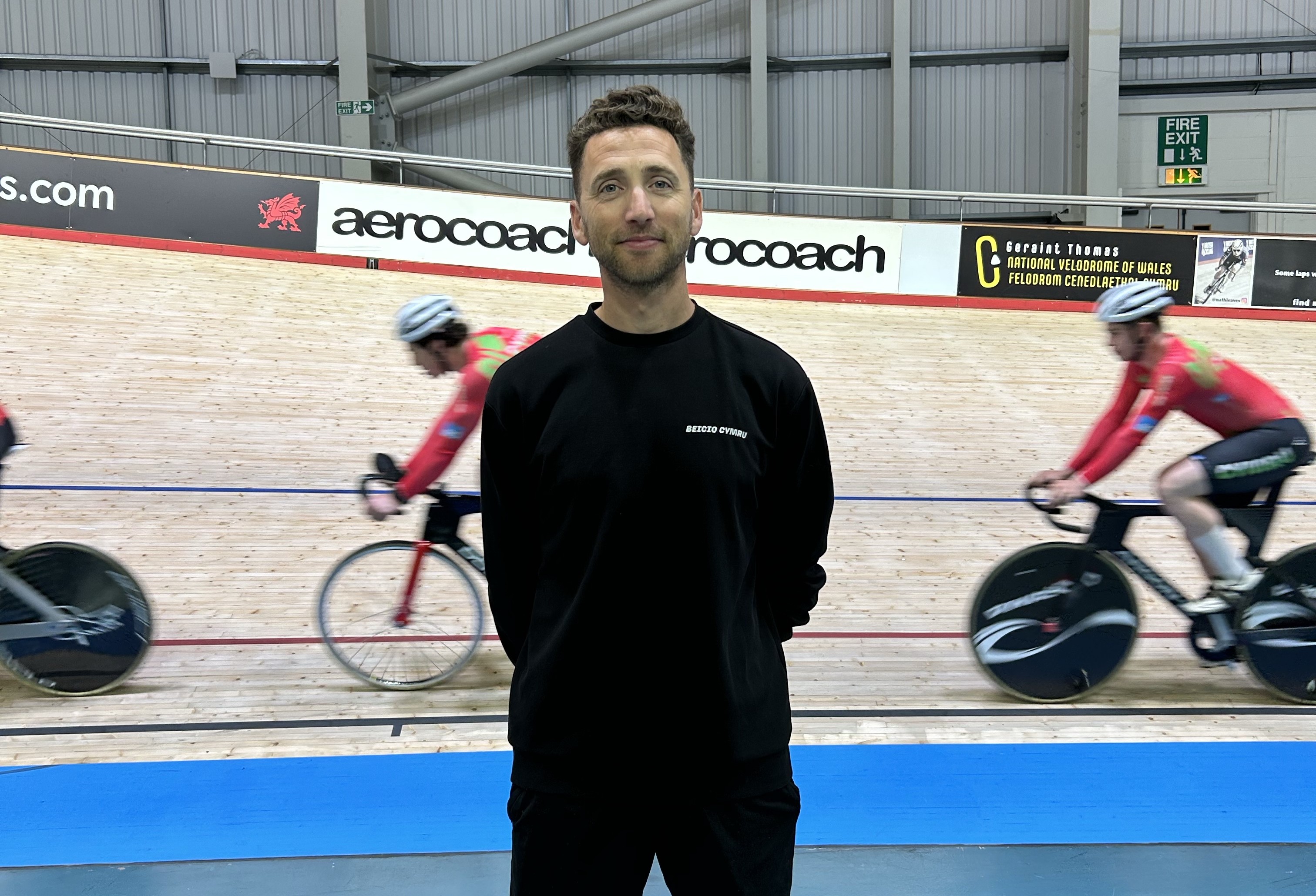 Rob stands on the infield of the Geraint Thomas National Velodrome of Wales while track cyclists ride past in the background during a training session.