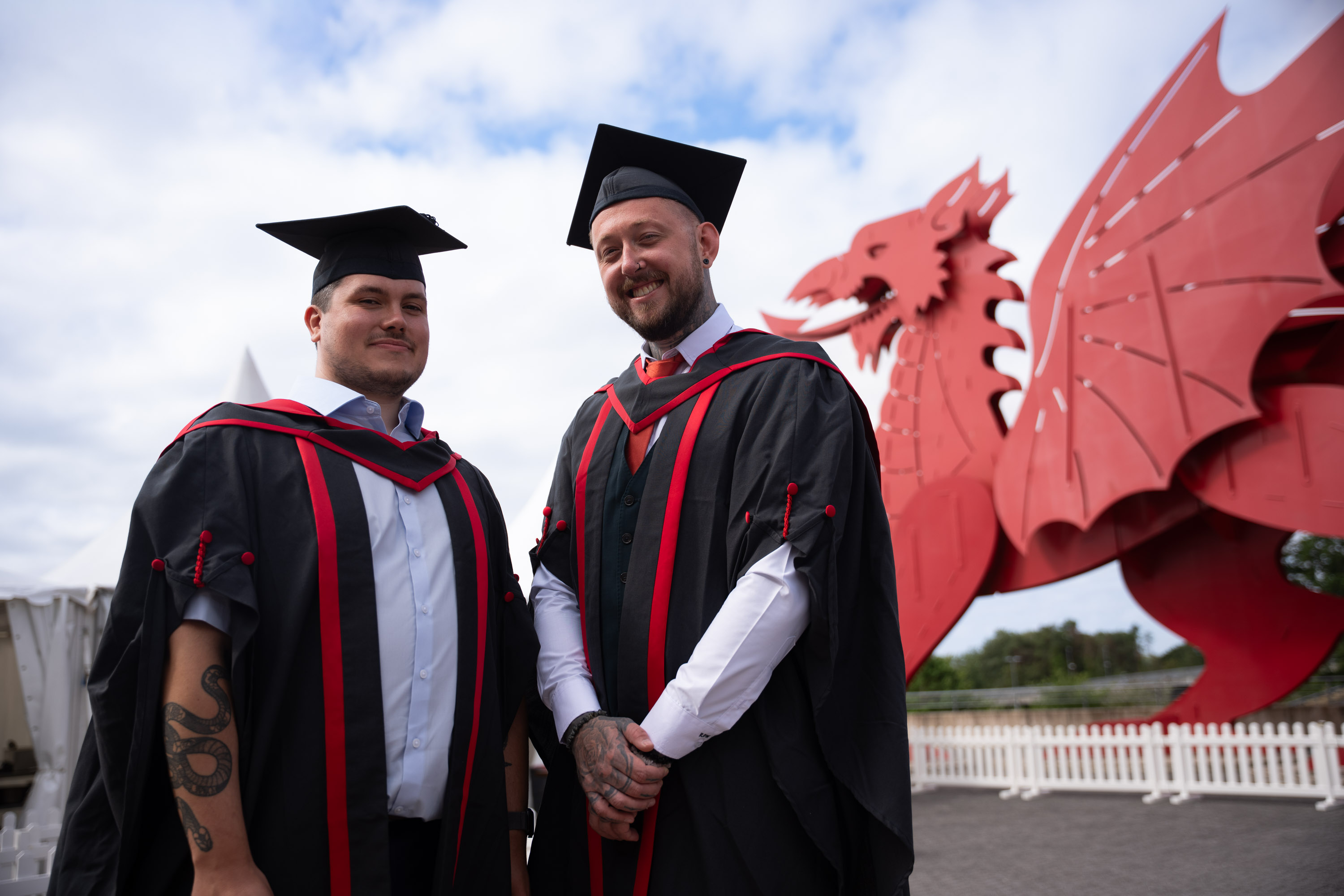 Ross Wills and Connor Jones, in their graduation gowns and mortarboards, in front of a giant dragon outside the ICC in Newport, Wales