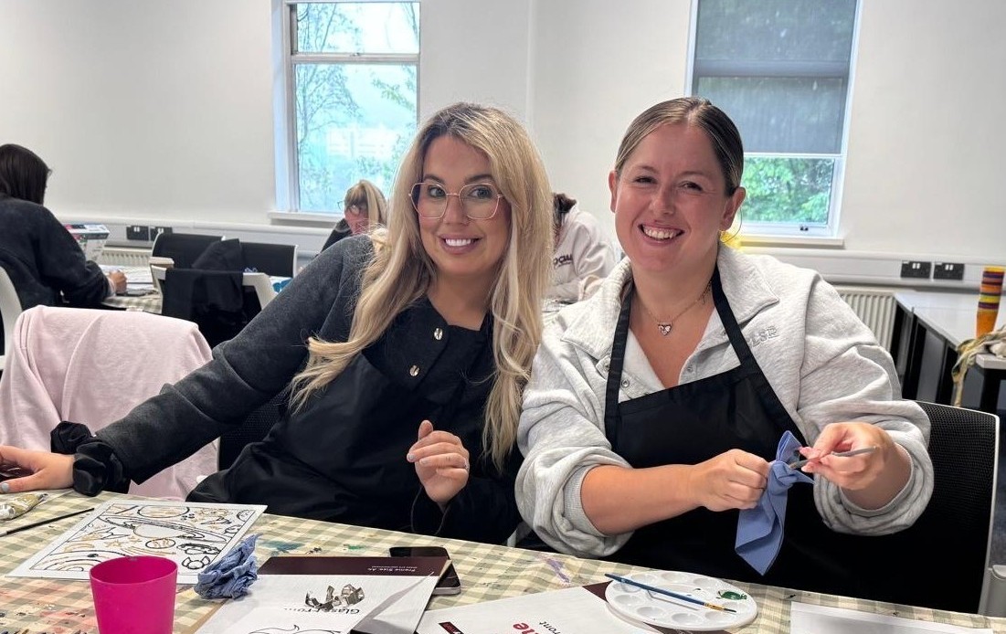 Two students sit together at a table during a wellbeing activity, there are paints and brushes on the table.